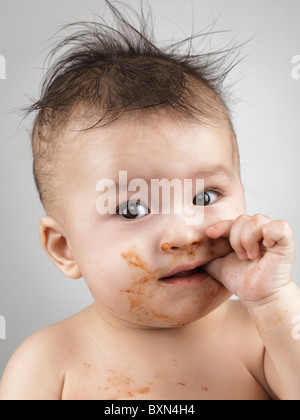 Künstlerische Porträt eines sieben Monate alten baby Boy mit strubbeligem Haar und verschmiert mit Essen Gesicht am Daumen lutscht