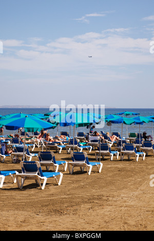 Touristen auf Liegestühlen und blauen Sonnenschirmen am Strand von Larnaca mit dem Flugzeug auf der Anfahrt zum Flughafen Stockfoto