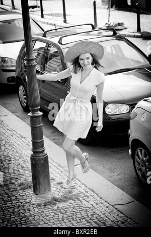 Schöne brünette Frau in weißem Kleid und Sonnenhut, die hinter Laternenpfahl auf der Kopfsteinpflasterstraße Prag Tschechien blickt Stockfoto