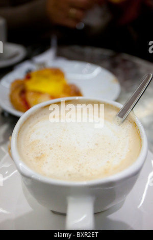 Nahaufnahme eines aufschäumenden Cappuccinos in einer weißen Tasse mit verschwommenem Gebäck im Hintergrund Stockfoto