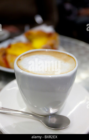 Nahaufnahme eines aufschäumenden Cappuccinos in einer weißen Tasse mit verschwommenem Gebäck im Hintergrund Stockfoto