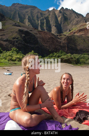 Zwei junge Frauen sitzen auf Kalalau Strand mit Bergen und Kajaks hinter Na Pali, Kauai Stockfoto