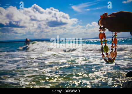 Eine Frau Hand Holding Perlenkette mit australischen Surfer Reiten ein Welle im Hintergrund, Tag der wunderschönen blauen Himmel mit Wolken Stockfoto