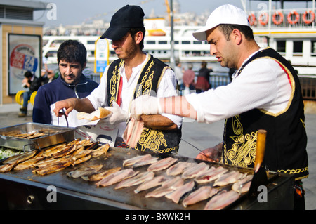 Balik Ekmek Fish Sandwich Street Food Eminonu Istanbul // ISTANBUL, Türkei – Ein Straßenverkäufer grillt frischen Fisch am Ufer von Eminonu in der Nähe der Galata-Brücke und bereitet das traditionelle balik ekmek (Fisch-Sandwich) zu. Der balik ekmek, der typischerweise aus gegrillter Makrele in Weißbrot mit Zwiebeln und Salat zubereitet wird, gehört zu den beliebtesten Street Food-Gerichten Istanbuls. Fischverkäufer am Wasser bedienen sich seit Generationen von schwimmenden Booten und Ständen entlang des Goldenen Horns. Der Stadtteil Eminonu dient als wichtiger Fährhafen und Handelsknotenpunkt zwischen Istanbuls europäischem und ASI Stockfoto