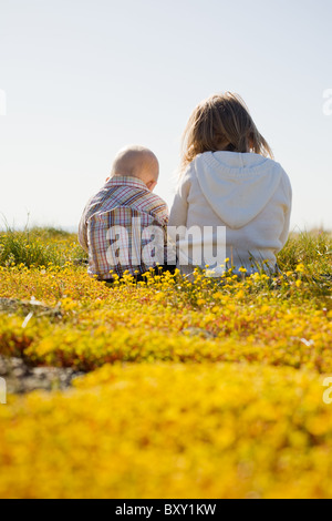 Ein Kleinkind Jungen und Mädchen sitzen mit dem Rücken zur Kamera in einem Feld von gelben Blüten. Stockfoto