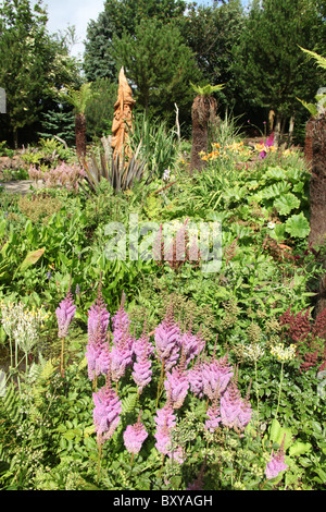 Mount Pleasant Gardens, England. Sommer-Ansicht der Astilben in voller Blüte mit einer Skulptur aus Holz Assistent im Hintergrund. Stockfoto