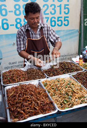 Mann, der gekochte Wanzen und Insekten verkauft, während er auf dem Chatuchak Market Bangkok Thailand eine Zigarette raucht Stockfoto