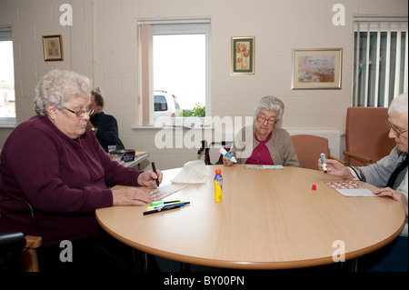 Drei ältere Freunde sitzen an Tisch im Tageszentrum Bingo zusammen zu spielen. Männliche Bingoanrufer im Hintergrund. Stockfoto