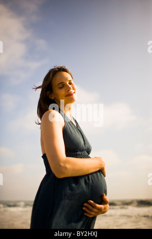 Eine schwangere Frau in einem schwarzen Halfter Kleid posiert für ein Porträt an einem sonnigen Tag am Strand. Stockfoto