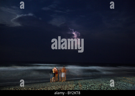 Menschen spielen am Strand in der Nacht während Blitze über den Ozean. Stockfoto