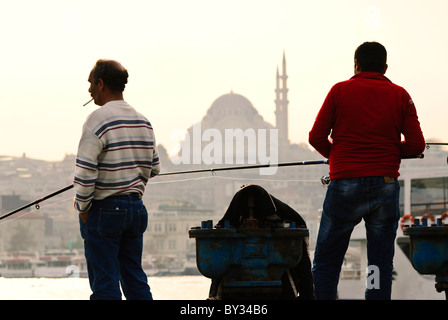 ISTANBUL, Türkei / Türkiye – Fischer werfen ihre Linien vom Ufer in der Nähe des Karakoy Fischmarktes entlang des Goldenen Horns. Die historische Galata-Brücke, ein beliebter Angelplatz, verbindet die Viertel Karakoy und Eminonu. Die berühmte Suleymaniye-Moschee, die 1557 während des Osmanischen Reiches fertiggestellt wurde, dominiert die Skyline. Stockfoto