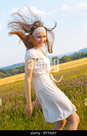 Fröhliche junge Frau, die sich mit Haaren dreht, die im Wind fliegen, in einem weißen Kleid auf einer Wildblumenwiese an einem sonnigen Sommertag in Tschechien Stockfoto