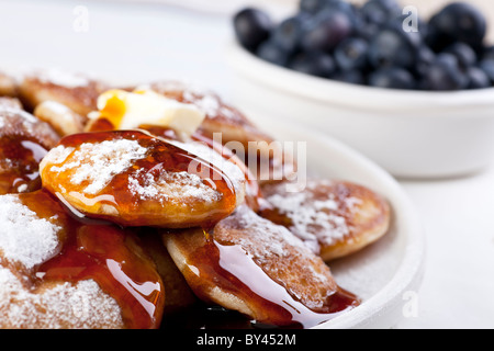 Holländische Mini-Pfannkuchen, oder Poffertjes mit Butter, Sirup und Puderzucker. Frische Heidelbeeren im Hintergrund unscharf. Stockfoto