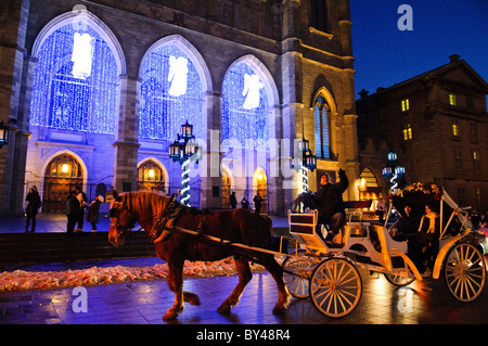 MONTREAL, Kanada – Eine Pferdekutsche mit Touristen fährt an der Vorderseite der Basilika Notre Dame in der Altstadt von Montreal vorbei, beleuchtet mit Engelslichtern, die den Eingang zur Weihnachtszeit schmücken. Die Basilika, bekannt für ihre neugotische Architektur, ist eines der berühmtesten Wahrzeichen der Stadt. Die saisonale Beleuchtung verstärkt die festliche Atmosphäre und zieht Besucher während der Feiertage an dieser historischen Stätte an. Stockfoto