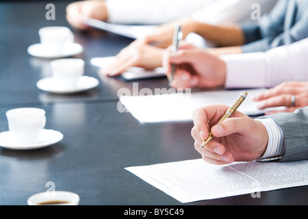 Nahaufnahme der männlichen Hand mit Kugelschreiber über Geschäftsdokument auf Hintergrund von Menschenhand mit Tassen Kaffee in der Nähe von Stockfoto