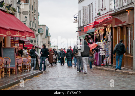 Montmartre Cobblestone Street Cafe Szene mit Menschen Paris France // PARIS, Frankreich – Eine kopfsteingepflasterte Straße in Montmartre bietet traditionelle Cafés mit roten Markisen und historischen Gebäuden, die von Fußgängern bevölkert werden. Dieses berühmte Viertel im 18. Arrondissement von Paris ist bekannt für sein künstlerisches Erbe und seine charmanten, verwinkelten Straßen. Montmartre war historisch ein Drehkreuz für Künstler wie Pablo Picasso und Vincent van Gogh, was zu seinem einzigartigen Bohemiencharakter beitrug. Heute zieht die einzigartige Atmosphäre und die malerische Umgebung zahlreiche Besucher und Kunstliebhaber an Stockfoto