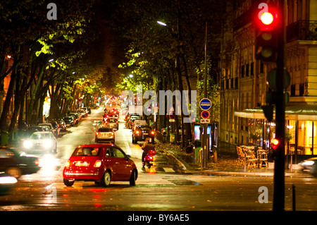 Paris Baum gesäumte Avenue nasser Straßenverkehr Abenddämmerung Frankreich // PARIS, Frankreich — nasse Straßen im 7. Arrondissement von Paris werden in der Abenddämmerung beleuchtet. Auf einer von Bäumen gesäumten Allee bewegen sich Fahrzeuge, deren Lichter auf dem feuchten Gehweg reflektieren. Dadurch entsteht eine leuchtende urbane Szene, die charakteristisch für das abendliche Ambiente der Stadt ist. Das Viertel liegt in der Nähe des berühmten Eiffelturms, einem weltberühmten Wahrzeichen der französischen Hauptstadt. Das 7. Arrondissement ist ein zentrales und elegantes Viertel innerhalb der Region Ile-de-France, bekannt für seine großartige Architektur und Regierungsgebäude. Stockfoto
