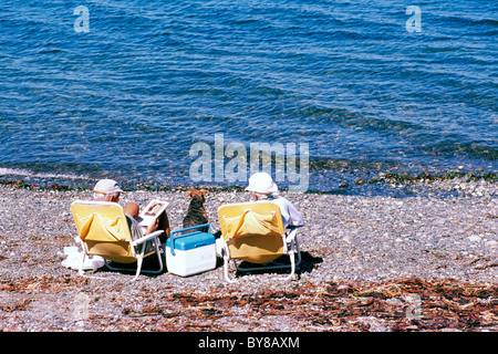 Ältere Paare mit Hund sitzen auf Stühlen und Sonnenbaden - Qualicum Beach, BC, Vancouver Island, British Columbia, Kanada Stockfoto