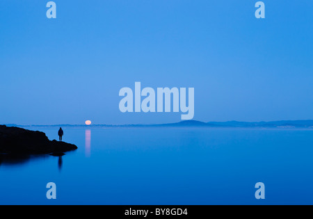 Eine Frau steht auf einer Küstenlinie beobachten die Einstellung Vollmond über Haro Strait und Vancouver Island, San Juan Island, WA, USA. Stockfoto