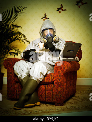 Mann sitzt in einem Sessel in einem inländischen Vorortlage mit seinem Laptop und eine Tasse Tee in einem thermonuklearen Anzug Stockfoto