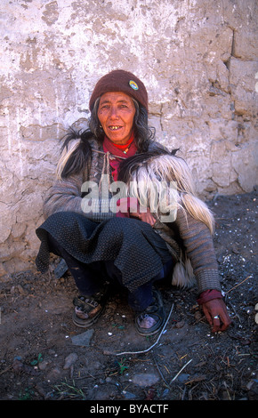 Ältere Frau tragen traditionelle Kleidung mit einem Ziegenfell über seine Schultern, sitzen auf dem Boden vor ihrem Haus, Zanskar Stockfoto