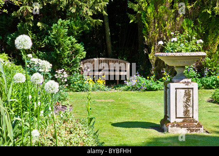 Eine kleine Liegewiese in einem englischen Landhaus-Garten im Sommer mit einer Holzbank in einer schattigen Hecke und einem Stein Pflanzer auf einem Sockel. Stockfoto