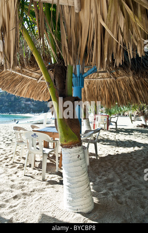 Tischen & Stühlen für Mahlzeiten im Freien, im Schatten der Sonnenschirme Palapa & Palmen am sonnigen Strand von Puerto Angel Oaxaca Zustand dargelegt Stockfoto