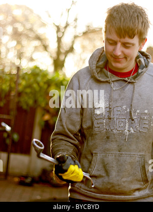 Einen jungen kaukasischen männlichen Mechaniker greifen einen Schraubenschlüssel, trägt ein paar schützende Rigger Handschuhe und ein Hoodie. Stockfoto