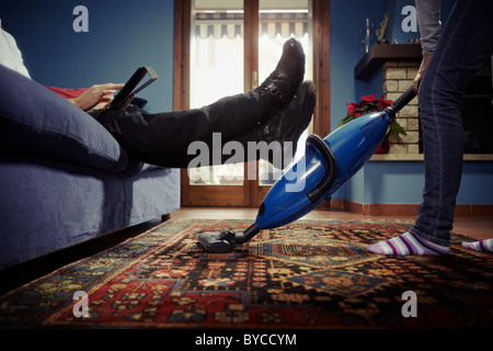 Kaukasische Heterosexuelles Paar, mit Frau tun Hausarbeit mit Staubsauger auf Teppich und faulen Mannes auf Sofa mit Tablet-pc. Stockfoto