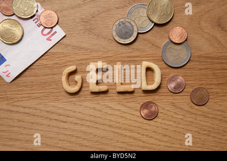 Geld, Deutsch für Geld, Wort, geschrieben mit Keksen, neben Münzen und Banknoten Stockfoto