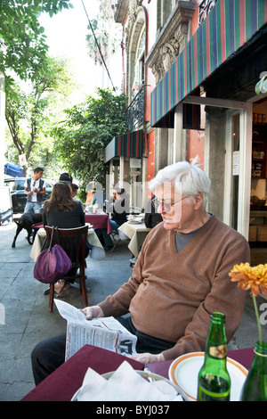 hochrangiger amerikanische Mann sitzt lesen Zeitung in hübschen Straßencafé mit Sitzgelegenheiten im Freien auf begrünten Seitenstraße Roma-Viertel Stockfoto