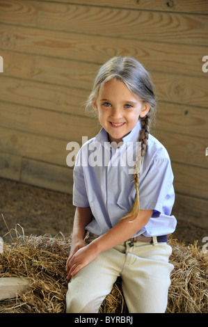 Kleine junge Mädchen sitzen auf einem Heuballen in einem Stall.  Das Mädchen hat lange blonde Haare, die geflochten ist. Stockfoto