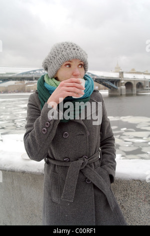 Die weißen gefrorenen Mädchen in einem blauen Schal, eine graue Kappe und einen Mantel trinkt heißen Tee aus einer Tasse auf Pushkinsky Kai, Moskau, Russland Stockfoto