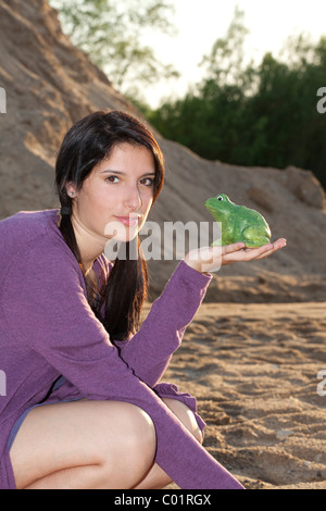Junge Frau hocken und halten einen grünen Keramik Frosch vor einer Sanddüne Stockfoto