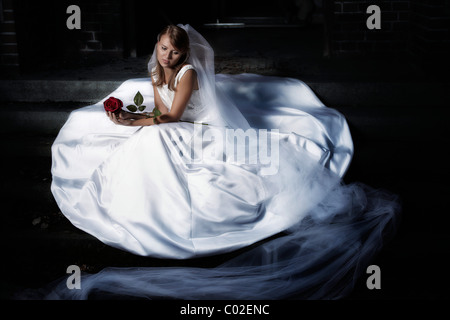 Junge blonde Frau in einem Hochzeitskleid und Schleier, sitzt auf einer Treppe in innerstädtischer Lage und hielt eine rote Rose in der hand Stockfoto