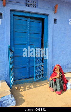 Ältere indische Frau trägt einen roten Sari sitzen auf dem Boden vor einer blau lackierten Tür, Thar-Wüste, Rajasthan, Indien Stockfoto