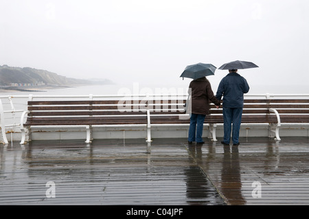 Reifen Sie, älteres Paar im Regen am Meer Pier stehen Stockfoto