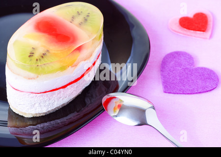 Nahaufnahme von Kuchen mit Gelee und Früchte auf einem schwarzen Teller mit Valentinstag Herz geformt Dekorationen auf lila Hintergrund Stockfoto
