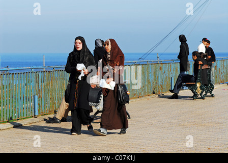 Algerien, Algier, Seitenansicht des Mädchen das Tragen der Burka zu Fuß durch Geländer während Paare sitzen im Hintergrund auf dem Seeweg Stockfoto