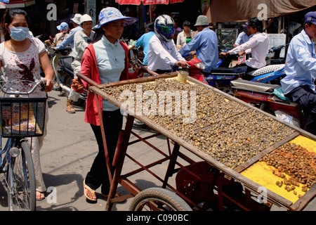 Eine Frau Lebensmittel Hersteller drängt einen Wagen beladen mit Schnecken auf einer belebten Straße in Phnom Penh, Kambodscha. Stockfoto