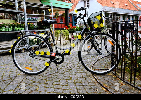 Ein Damen Fahrrad dekoriert mit Narzissen Aufkleber links in einem Fahrradständer außerhalb Geschäfte auf einer gepflasterten Straße in Ronne Bornholm Stockfoto