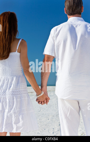 Rückansicht des Mannes & Frau romantisch zu zweit in weißen Kleidern, die Hand in Hand auf einem einsamen tropischen Strand mit strahlend blauem Himmel Stockfoto