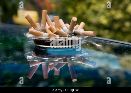 EIN VOLLER ASCHENBECHER ZIGARETTE ZIGARETTENKIPPEN AUF EINEM GLASTISCH IM GARTEN Stockfoto