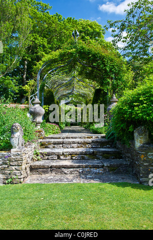 Steinstufen führen zu einer Pergola über einen gepflasterten Pfad in einem englischen Landhaus-Garten mit einem hölzernen Sitz am Ende. Stockfoto