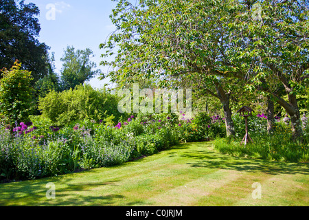 Blumenbeete oder Betten rund um einen gestreiften Rasen in einem englischen Landhaus-Garten im Sommer mit hölzernen Vogel Tisch unter Bäumen. Stockfoto