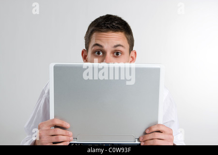 Ein junger Mann hält einen Laptop-Computer eng und lugt über die Oberseite mit hochgezogenen Augenbrauen. Stockfoto