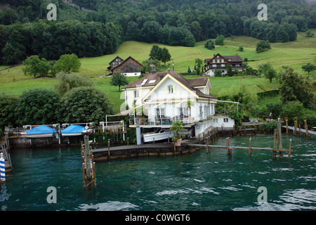 Ein Haus, Pier und Boote am Ufer des Vierwaldstättersees in der Schweiz. Das Haus liegt auf einem grünen Hügel mit anderen Häusern. Stockfoto