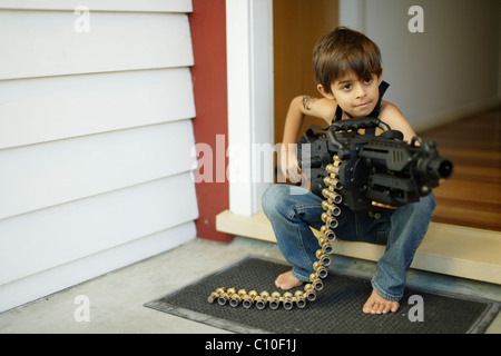 Siebenjähriger Junge sitzt vor Haustür hält Spielzeug Maschinengewehr Stockfoto