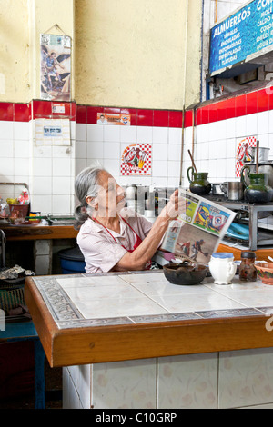 elegante ältere einheimische Frau Koch sitzt in ihrem makellosen Fliesen Markt Alkoven Lektüre offen Zeitung am Nachmittag Flaute Oaxaca Stockfoto