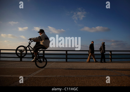 Ein älteres Ehepaar zu Fuß entlang der Strandpromenade, während ein Junge auf seinem Mountainbike, Alabaster Küste, Normandie Tricks ausführt Stockfoto